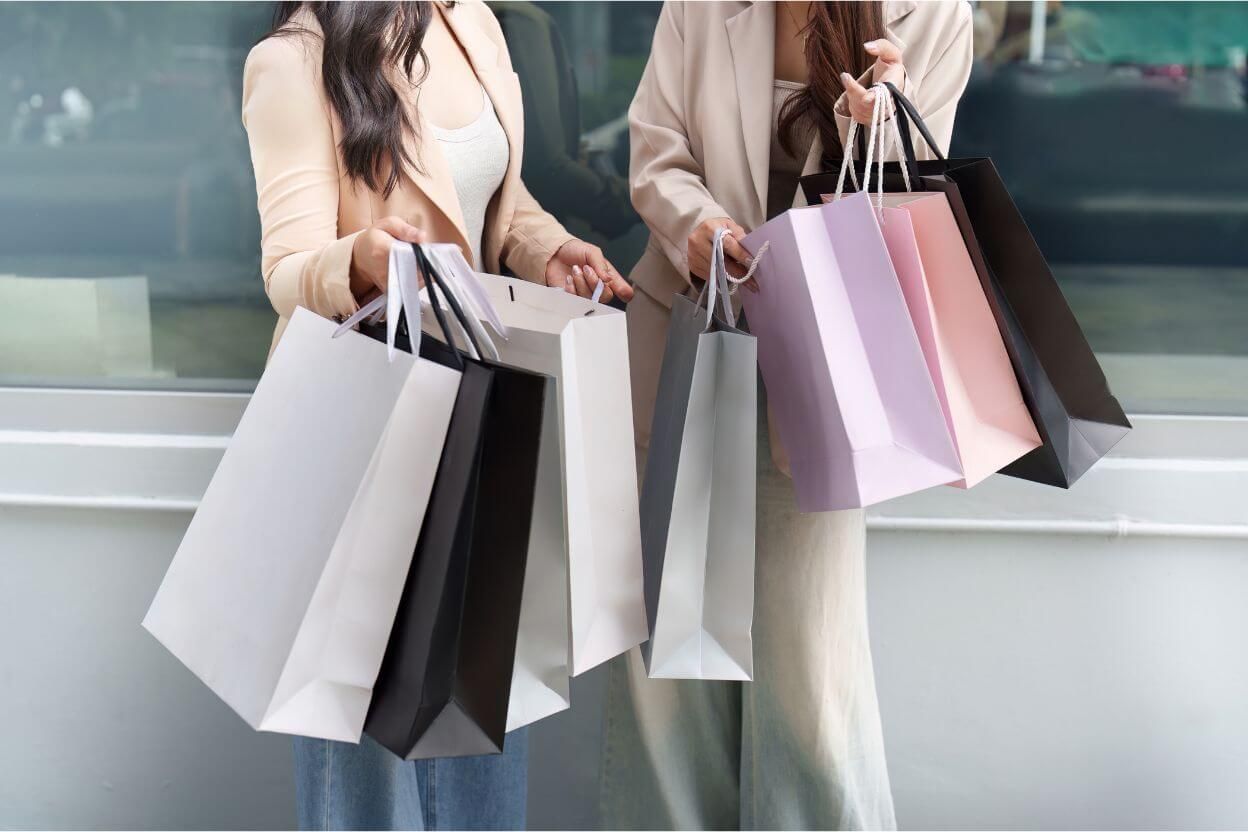Divas sievietes ar vairākām iepirkumu somām - simbols impulsīviem pirkumiem un naudas tērēšanas ieradumiem.
